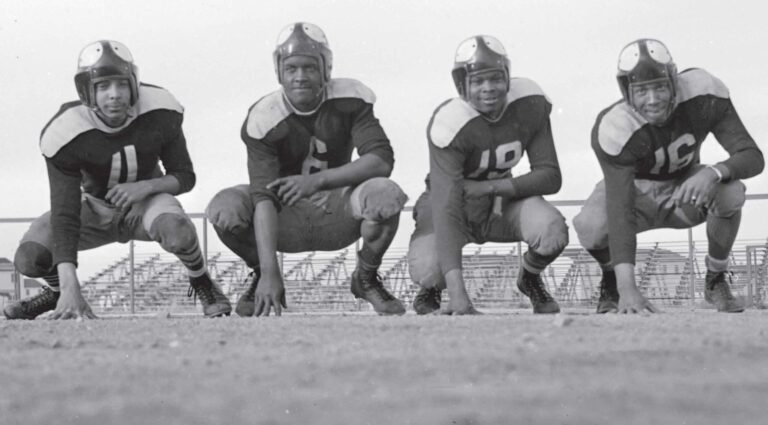 An image of four young Black men in 1940s football uniforms, crouching in front of bleachers.