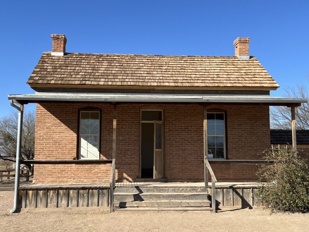 Camera picture taken in January 2025 of the front of the Bryce red brick house with 4 step wooden porch. The rectangular house is one story with the front door centered between two windows with two chimneys on each end of the house.