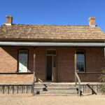 Camera picture taken in January 2025 of the front of the Bryce red brick house with 4 step wooden porch. The rectangular house is one story with the front door centered between two windows with two chimneys on each end of the house.