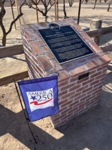 Photo showing the 4 foot red brick marker with description plaques pertaining to the home and the bricks. The plaques are placed on the top and front of the marker. To the left of the marker is a yard flag which reads “America 250 Daughters of the American Revolution”.