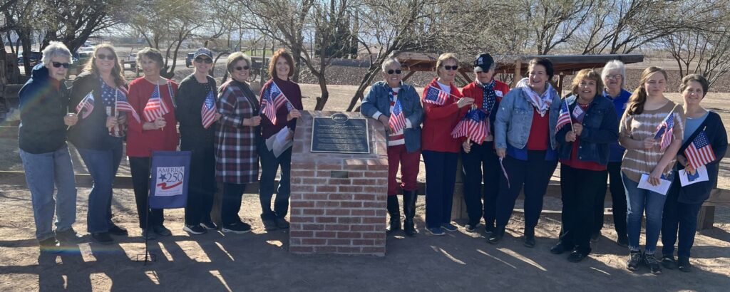 A group of 14 ladies from the National Society of Daughters of the American Revolution Gila Valley Chapter holding American flags while standing on both sides of the 4 foot brick marker. In the ground is an America 250 yard flag. The photo was taken at the unveiling of the Bryce Brick House marker on January 18, 2025.